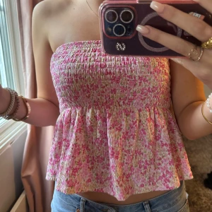 Blommig bandeau tubtopp i rosa och vit - Supersöt bandeau tubtopp med smockad överdel och volangig nederdel. Toppens tyg är blommigt i rosa, vitt och lite grönt, perfekt för sommarens alla tillfällen. Sitter bekvämt och ärmlös för en luftig känsla. Passar till jeans eller kjol för en fräsch look.