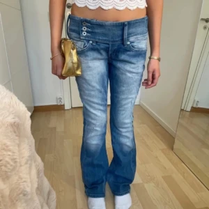 Blåa lågmidjade bootcut jeans från Diesel - Säljer ett par blåa jeans från Diesel med låg midja och bootcut passform. Jeansen har en unik knappstängning i sidan och snygga slitningar framtill. Bakfickorna har broderade detaljer och en stor Diesel-logga på bakre linningen. Perfekta för dig som gillar retrovibbar.