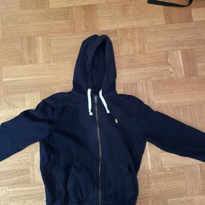 Marinblå ziphoodie från Polo Ralph Lauren - Säljer en marinblå hoodie från Polo Ralph Lauren med dragkedja och vit snörning i huvan. Klassisk gul logga på bröstet och två fickor framtill. Perfekt för dig som gillar stilrena och bekväma plagg. Mycket bra skick och mycket lågt pris!👊