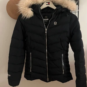 Svart pufferjacka 8848 Altitude med fluff runt huvan - Svart pufferjacka från 8848 Altitude med snygg fluff runt på huvan och silvriga dragkedjor. Jackan har två fickor med dragkedja och passformad i midjan. 