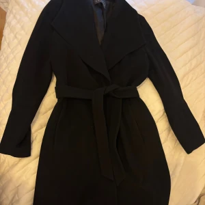 Svart kappa med bälte från Esprit - Stilren svart kappa från Esprit i storlek S med bred krage och knytskärp i midjan. Långärmad modell i klassisk passform, perfekt för kyligare dagar. Kappan har en elegant siluett och är tillverkad i ett mjukt material som känns lyxigt.