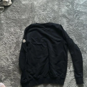 Svart sweatshirt från Moncler - Svart sweatshirt från Moncler med rund hals och ribbade muddar. Klassisk Moncler-logga på vänster ärm och diskret broderad detalj på bröstet. Perfekt för dig som gillar stilrena och exklusiva plagg med skön passform.