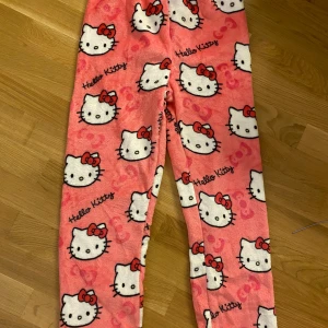 Hello Kitty rosa mjukisbyxor - Supermysiga rosa mjukisbyxor med Hello Kitty-motiv över hela byxorna. Byxorna är i fleece och har ett gulligt tryck med Hello Kittys ansikte och röda rosetter. Lite små i storlek 