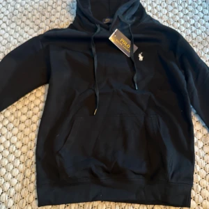 Svart hoodie från Polo Ralph Lauren - Svart hoodie från Polo Ralph Lauren med klassisk huva och snörning. Tröjan har en broderad logga på bröstet och en stor magficka framtill. Perfekt för dig som gillar stilren och enkel design med premiumkänsla. Storlek M men sitter mer som en S