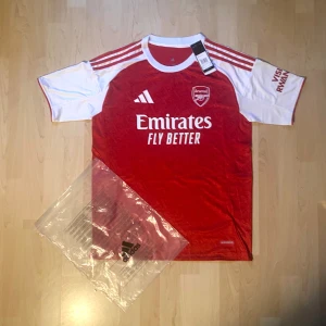 Arsenal Gyökeres 14 matchtröja Adidas - Röd och vit Arsenal fotbollströja från Adidas med Gyökeres och nummer 14 på ryggen. Klassisk design med korta ärmar, klubbmärke och sponsortryck framtill. Tillverkad i lätt och ventilerande material, perfekt för match eller träning.