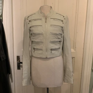 Ljusgrå croppad skinnjacka med dragkedjor - Säljer en ljusgrå croppad skinnjacka med coola silvriga dragkedjor framtill och på ärmsluten. Jackan har en rak, kort passform och ståkrage. Perfekt för dig som vill sticka ut med en edgy look.