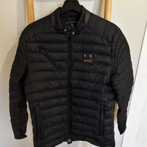 Svart du jacka från Morris *M* - Svart pufferjacka från Morris med quiltad design och snygga detaljer i skinn vid kragen. Jackan har dragkedja framtill, två sidofickor med dragkedja och en bröstficka med sned dragkedja. Märkespatch på bröstet och stilren look. Perfekt för hösten.