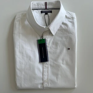 Vit skjorta från Tommy Hilfiger - Vit klassisk skjorta från Tommy Hilfiger med stilren design och diskret logga broderad på bröstet. Skjortan har lång ärm, knappar framtill och är tillverkad i mjuk bomull. Perfekt för dig som gillar clean och tidlös stil. Skjortan är oanvänd med saknar prislapp.