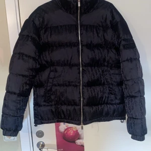 Svart pufferjacka från Dior - Snygg svart pufferjacka från Dior med diskret monogrammönster över hela jackan. Jackan har dragkedja framtill, hög krage och två fickor. Materialet är glansigt och känns lyxigt, perfekt för kalla dagar när du vill sticka ut med stil.