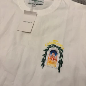 Vit t-shirt från Casablanca med tryck - Vit t-shirt från Casablanca i 100% bomull med färgglatt tryck av en byggnad och gröna blad både fram och bak. Klassisk passform och rund halsringning, perfekt för dig som gillar stilrena men ändå unika plagg.