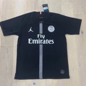 Svart PSG fotbollströja Jordan x Fly Emirates - Snygg svart PSG fotbollströja med korta ärmar, tillverkad i lätt och ventilerande material. Framsidan har Fly Emirates-tryck, PSG-logga och Jordan-logga samt en grå vertikal rand. Perfekt för dig som vill sticka ut på planen eller på läktaren.