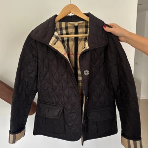 Svart quiltad jacka från Burberry - Stilren svart quiltad jacka från Burberry med klassiskt rutigt foder i beige, svart och rött. Jackan har stora fickor framtill och stängs med knappar. Perfekt för dig som vill ha en tidlös och snygg look med lyxig känsla.