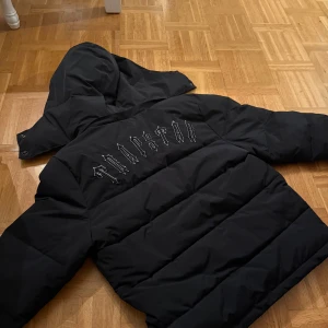 Svart pufferjacka från Trapstar - Säljer en svart pufferjacka från Trapstar med stor logga på ryggen och patch på bröstet. Jackan har avtagbar huva, dragkedja framtill och två fickor. Perfekt för kalla dagar.