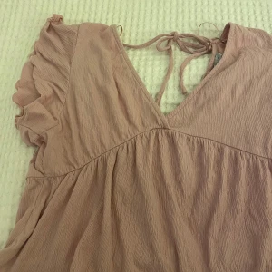 Ljusrosa volangtopp från 157 - Supersöt ljusrosa topp från 157 med volangärmar, v-ringning och knytning i nacken. Tillverkad i ett lätt, krinklat tyg som ger en avslappnad känsla. Perfekt för varma dagar när du vill känna dig både bekväm och stylish💞