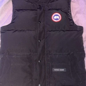 Svart dunväst från Canada Goose - Svart dunväst från Canada Goose med klassisk logga på bröstet och två stora fickor framtill. Västen har tryckknappar och en hög krage som ger extra värme. Perfekt för kalla dagar och har en stilren, enkel design.