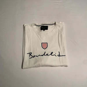 Vit långärmad tröja från Bondelid - Vit långärmad tröja från Bondelid med broderad logga och rödvit sköld på bröstet. Tröjan har rund halsringning och är tillverkad i mjuk bomull. Perfekt för en clean och stilren look.