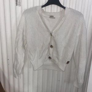 Vit stickad kofta med knappar - Mysig vit stickad kofta från Lindex med stora bruna knappar framtill. Koftan har en avslappnad passform, ribbade muddar och lång ärm. Perfekt att slänga på sig över en t-shirt eller linne för en chill look.