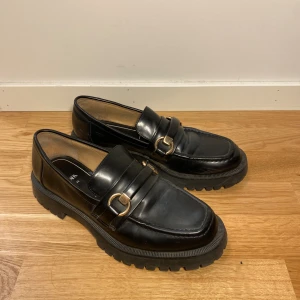 Svarta loafers från Zara med gulddetaljer - Säljer ett par svarta loafers från Zara i skinnimitation med chunky sula och snygga guldfärgade spännen framtill. Skorna har fyrkantig tå och en klassisk look som passar till många outfits.🖤👔