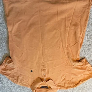 Orange pikétröja från Morris - Snygg orange pikétröja från Morris med klassisk krage, två knappar och broderad blå logga på bröstet. Tillverkad i mjuk bomullspiké som känns skön mot huden. Perfekt för dig som gillar stilren och avslappnad look.