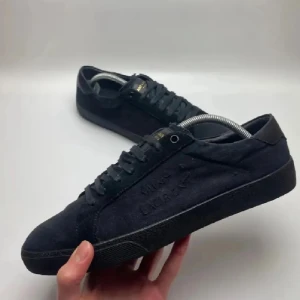 Saint Laurent SL/06 Black - Snygga svarta sneakers från Saint Laurent med diskret logga på sidan. Skorna har ovandel i mocka och detaljer i skinn, samt svarta snören och platt sula. Perfekta för dig som gillar stilrena och exklusiva sneakers med en lyxig känsla.