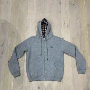 Burberry zip grå - Säljer en grå hoodie från Burberry med klassiskt rutigt foder i huvan. Tröjan har dragkedja framtill, snörning i huvan och två fickor. Perfekt för dig som vill ha en stilren och bekväm look med en touch av lyx.