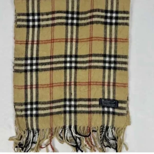 Rutig halsduk från Burberry i ull - Snygg rutig halsduk från Burberry i klassiska färger. Den har fransar i ändarna och är perfekt för att hålla dig varm och stilren under kyliga dagar. Halsduken är som pricken över I:t på din outfit. Skriv vid funderingar!