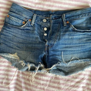 Levi's blå jeansshorts med fransig kant - Säljer ett par klassiska blå jeansshorts från Levi's med fransig och rå kant, fem fickor och knappgylf. Shortsen har hög midja och den ikoniska Levi's-lappen baktill. Perfekta för sommarens chill dagar och festivaler.