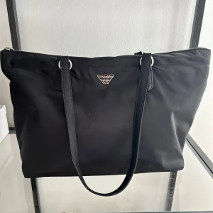 Svart axelväska från Emporio Armani - Stilren svart axelväska från Emporio Armani med silverfärgad logga framtill. Väskan har långa axelband och är tillverkad i ett slätt syntetmaterial. Invändigt finns ett rymligt fack med dragkedja och diskret mönstrat foder med märkets logga.