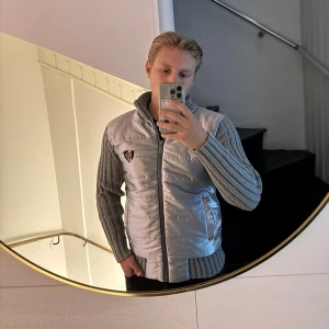 Grå cardigan från Tommy Hilfiger  - Säljer en grå dunväst från Tommy Hilfiger . Jackan har quiltad framsida i glansigt material, ribbstickad rygg och ärmar samt hög krage. Snyggt Hilfiger-emblem på bröstet och dragkedja framtill. Perfekt för lager-på-lager och streetstyle. Passar S/M