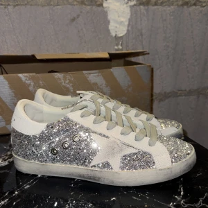 Glittriga golden goose med stjärna - Säljer ett par riktigt snygga sneakers med silverglittrig ovandel och en ikonisk stjärndetalj på sidan. Skorna har grå snörning, rund tå och detaljer i mocka. Perfekta för dig som vill sticka ut med din stil och älskar glitter.