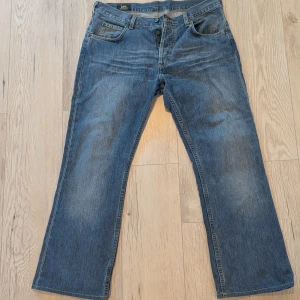 Blå bootcut jeans från Lee Denver - Säljer ett par klassiska blå jeans från Lee, modell Denver. Jeansen har bootcut passform, fem fickor och knäppning med Lee-logga på knapparna. Tillverkade i 100% bomull för skön känsla och slitstyrka. Perfekt för dig som gillar retrovibbar och bredare ben.