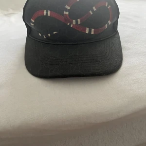 Svart Gucci keps ormen - Svart keps från Gucci med ikoniskt ormtryck i rött och vitt framtill. Justerbar rem baktill med röd och marinblå rand. Kepsen är äkta  storlek S 57cm