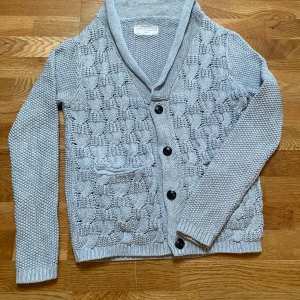Grå stickad cardigan från These Glory Days - Säljer en grå stickad kofta från These Glory Days i storlek medium. Koftan har snygg strukturstickning, knappar framtill, sjalkrage och en ficka på ena sidan. Perfekt för lager-på-lager och chillare dagar.