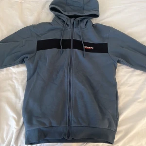 Blå hoodie från McKenzie med dragkedja - Snygg blå hoodie från McKenzie med svart band över bröstet och logga. Tröjan har hel dragkedja, huva med snörning och är fodrad med mjuk fleece på insidan. Perfekt för kyliga dagar och chill outfits.