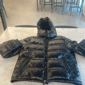 Svart Moncler pufferjacka med huva - Svart pufferjacka från Moncler med glansig finish och huva. Jackan har dragkedja framtill, två sidofickor med dragkedja och en ficka med Moncler-logga på ärmen. Tillverkad i polyester och dun för extra värme. Perfekt för kalla dagar.