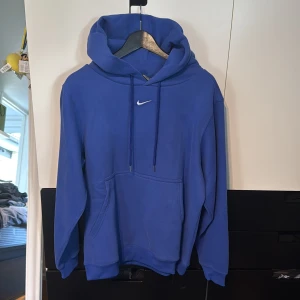Nike hoodie - Blå hoodie från Nike med klassisk logga broderad på bröstet. Tröjan har stor huva med snörning, magficka och ribbade muddar. Mjuk insida i fleece som gör den extra skön. Perfekt för chill eller träning, enkel och stilren design. Aldrig använd.