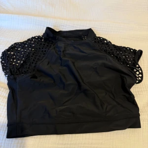 Svart croppad bad topp från Bik Bok - Säljer en svart croppad topp från Bik Bok med coola nätärmar och hög hals. Toppen är ärmlös med ett sportigt snitt och tillverkad i ett stretchigt syntetmaterial. Perfekt för dig som vill sticka ut med en edgy vibe.