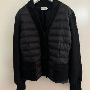 Svart puffer cardigan jacka Moncler S - Svart puffer cardigan jacka från Moncler i storlek S. Jackan har quiltad framsida i syntetmaterial och stickade ärmar och rygg i ull. Klassisk Moncler-logga på ärmen, ribbad krage och knäppning framtill. Två fickor med lock framtill ger en snygg och praktisk look.