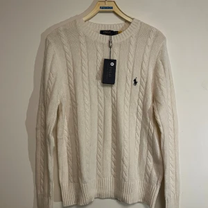 Vit kabelstickad tröja Polo Ralph Lauren - Kabelstickad vit tröja från Polo Ralph Lauren med klassisk rund halsringning och broderad logga på bröstet. Tröjan är långärmad och har ribbade muddar vid ärmslut och nederkant. Perfekt för dig som gillar stilren och tidlös design.