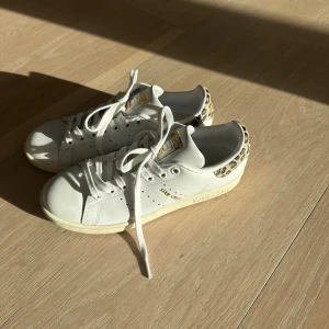 Adidas Stan Smith med leoparddetalj - Säljer ett par vita Adidas Stan Smith sneakers med snygga leopardmönstrade detaljer på hälen och guldfärgade loggor. Klassisk siluett med snörning, rund tå och platt sula. Skorna är i skinn och har en fräsch och trendig look som sticker ut.