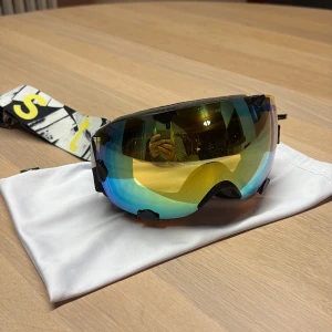 Barn Spegelglas skidglasögon med mönstrat band - Säljer ett par barn skidglasögon från Everest med spegelglas i blått och gult. Bandet är svartvitt med gula bokstäver och har ett coolt grafiskt mönster. Ramen är svart och glasögonen har en bred passform som täcker bra. Uppsakkata att de passar ett barn från 3-10år. Skyddspåse medföljer 