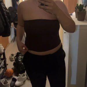 Mörkbrun bandeau tubtopp - Säljer en stilren mörkbrun bandeau tubtopp som sitter tight och har en enkel, rak form. Toppen är axelbandslös och passar perfekt för dig som gillar minimalistisk stil. Materialet känns mjukt och stretchigt mot huden.