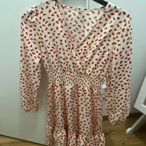 Vit och röd prickig omlottklänning - Supersöt kortklänning i vitt med röda prickar, omlottmodell och smock i midjan. Klänningen har långa ärmar med lätt puff och volang nertill. Perfekt för dig som gillar lekfulla mönster och en feminin stil.