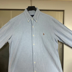 Ljusblå skjorta från Ralph Lauren - Klassisk ljusblå skjorta från Ralph Lauren med button-down krage och broderad logga på bröstet. Skjortan har långa ärmar och knäppning framtill. Perfekt för en clean och stilren look. Även försiktigt använd.