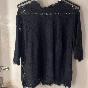 Svart spetsblus med blommönster - Svart blus i genombruten spets med stora blommor. Blusen har knapp i nacken och fina spetsdetaljer längs kanterna. Perfekt för dig som gillar romantisk och elegant stil.