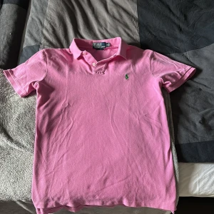  pikétröja från Ralph Lauren - Säljer en klassisk rosa pikétröja från Ralph Lauren Tröjan har den ikoniska gröna broderade loggan på bröstet.