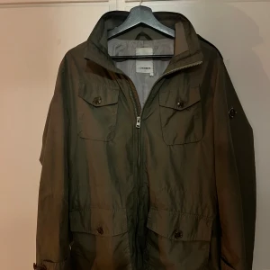 Grön vindjacka från J.Lindeberg - Snygg militärgrön vindjacka från J.Lindeberg med fyra stora fickor framtill och dragkedja hela vägen upp. Jackan har klassisk krage, axelklaffar och diskret logga på ärmen. Perfekt för dig som gillar stilren och funktionell design. 