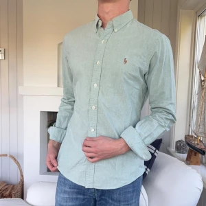 Ljusgrön skjorta från Polo Ralph Lauren - Snygg ljusgrön skjorta från Polo Ralph Lauren i slim fit-modell. Skjortan har klassisk button down-krage, broderad logga på bröstet och vita knappar framtill. Tillverkad i bomull och passar perfekt till jeans för en clean look.