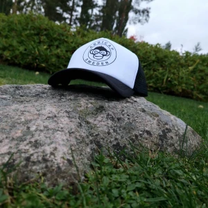 Svartvit truckerkeps från ABBICAP - Snygg truckerkeps från ABBICAP Sweden med svart nät baktill och vit frontpanel. Kepsen har ett tryckt apmotiv och rundad skärm. Perfekt för dig som vill ha en cool och avslappnad stil.