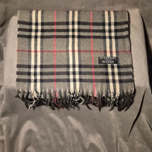 Rutig halsduk från Burberry i ull - Klassisk rutig halsduk från Burberry i 100% lammull. Färgerna är svart, grå, vit och röd med fransar i ändarna. Den ikoniska designen gör den till ett riktigt statement-plagg för kalla dagar. Perfekt accessoar för att lyfta din vinterstil.